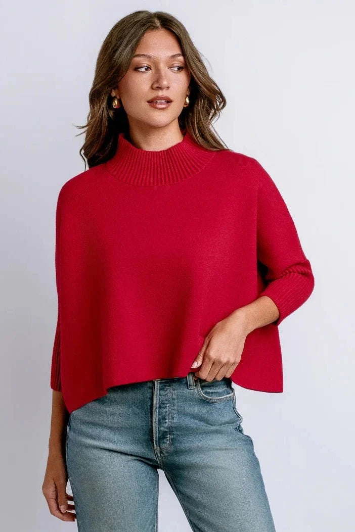 A woman wearing a bright red mock-neck knit sweater with three-quarter sleeves and blue jeans, standing against a light gray studio background.