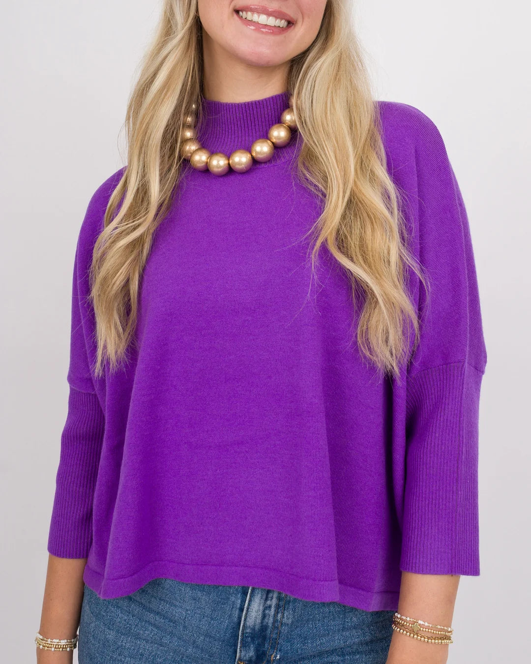 A woman wearing a bright purple knit mock-neck sweater with three-quarter sleeves, paired with a gold beaded necklace and blue jeans, against a light gray background.