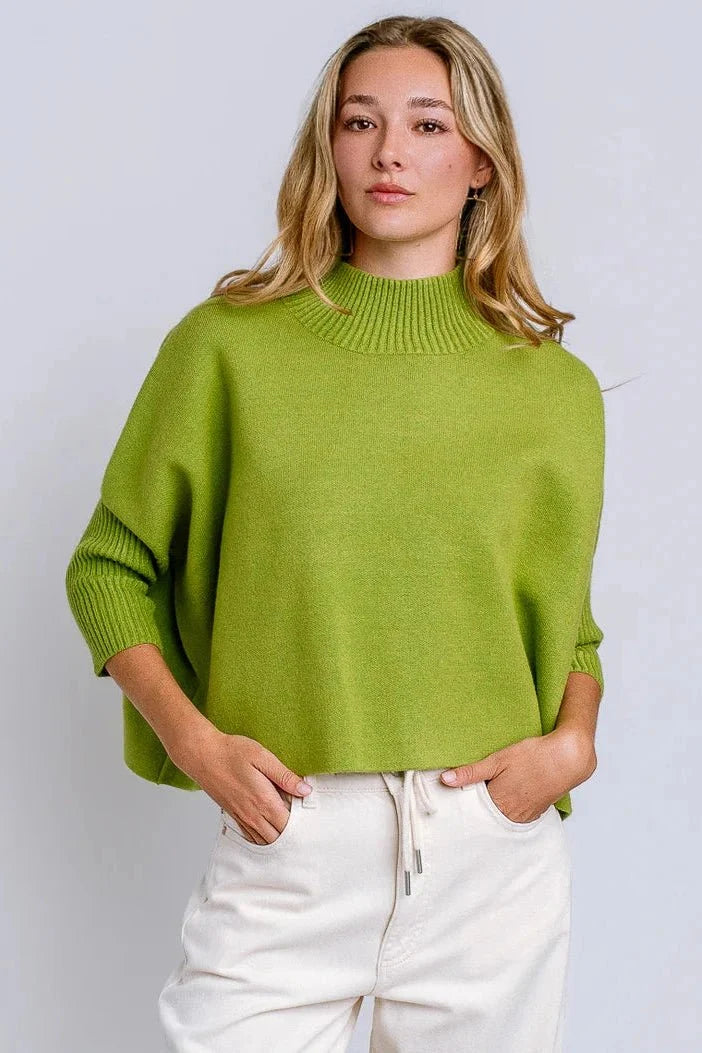 A woman wearing a bright lime green knit mock-neck sweater and white drawstring pants, standing against a light gray studio background.
