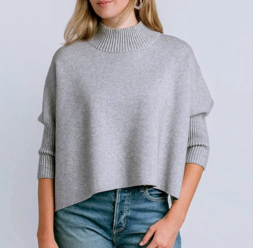 A woman wearing a light gray knit mock-neck sweater with ribbed three-quarter sleeves and drop shoulders, paired with blue jeans, against a plain light gray background.